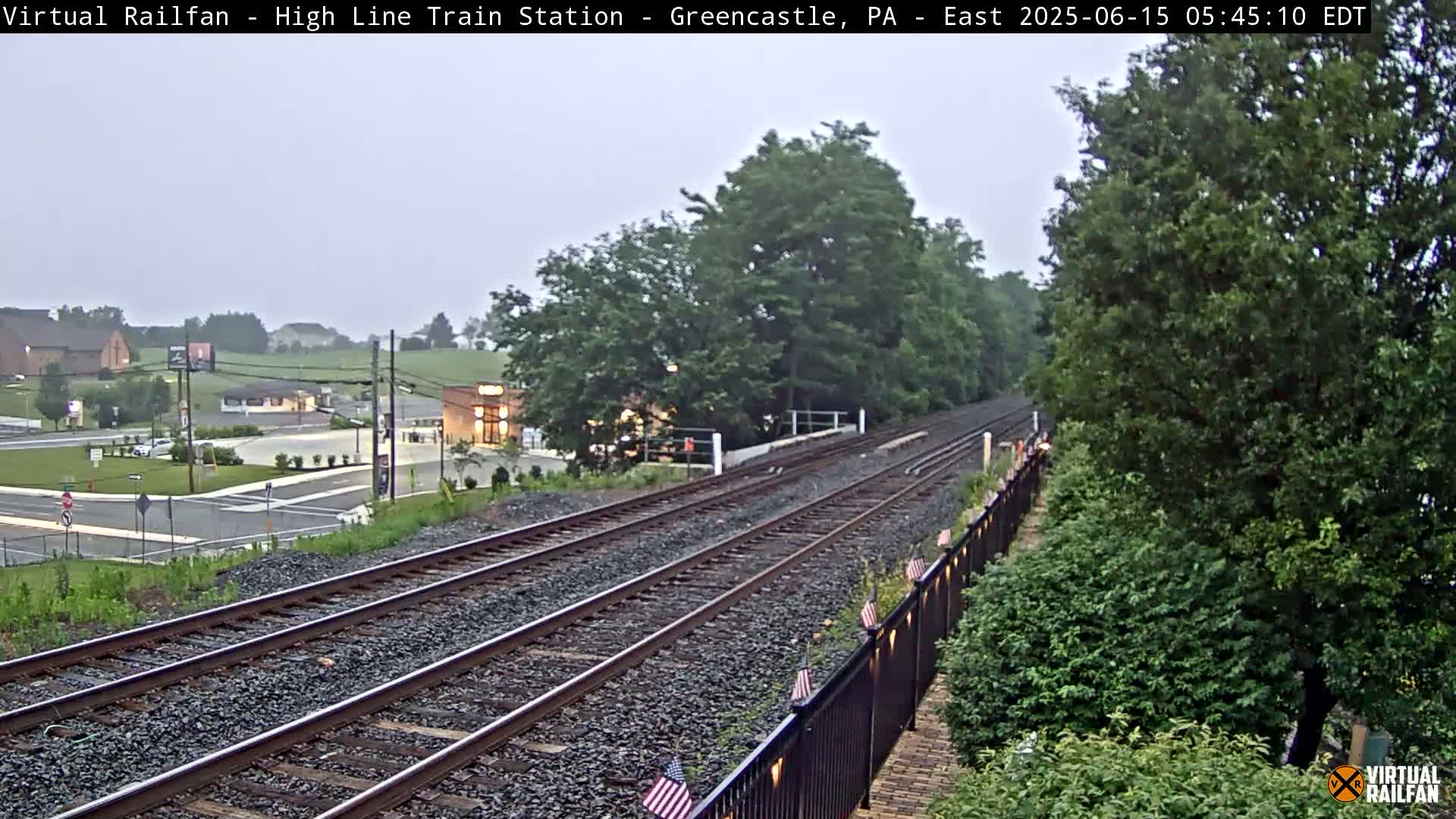 A view down a set of train tracks on an overcast day, with a small town and green foliage visible in the background.