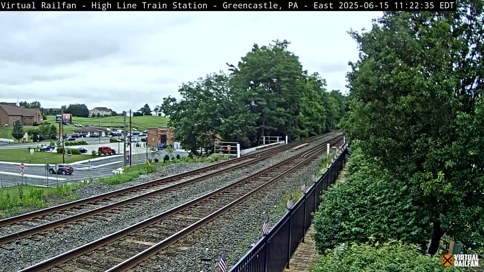 Two sets of train tracks extend into the distance, flanked by lush green vegetation and a black metal fence under a cloudy sky.