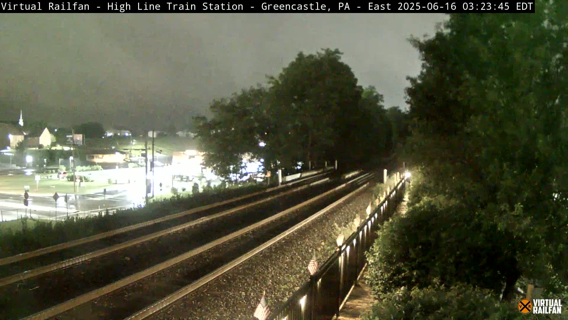 A nighttime view of a train track with a wet road and buildings visible in the background under an overcast sky.