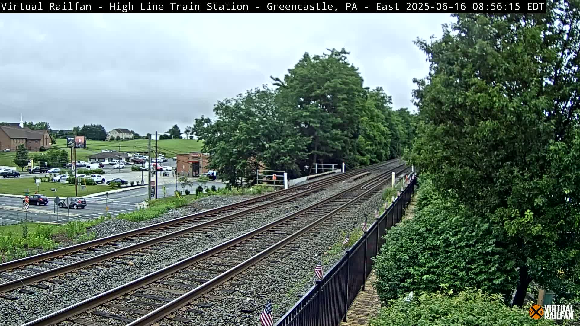 A view of a train track extending into the distance under an overcast sky, with green vegetation alongside and a parking lot and buildings visible in the background.