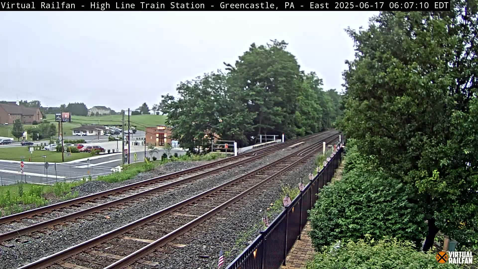 Two sets of train tracks extend into the distance, bordered by lush green vegetation and a black metal fence, under an overcast sky.