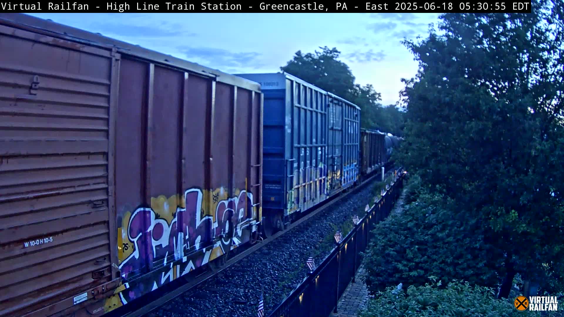 A freight train with graffiti-covered cars moves eastward along a track beside a line of trees under a twilight sky.