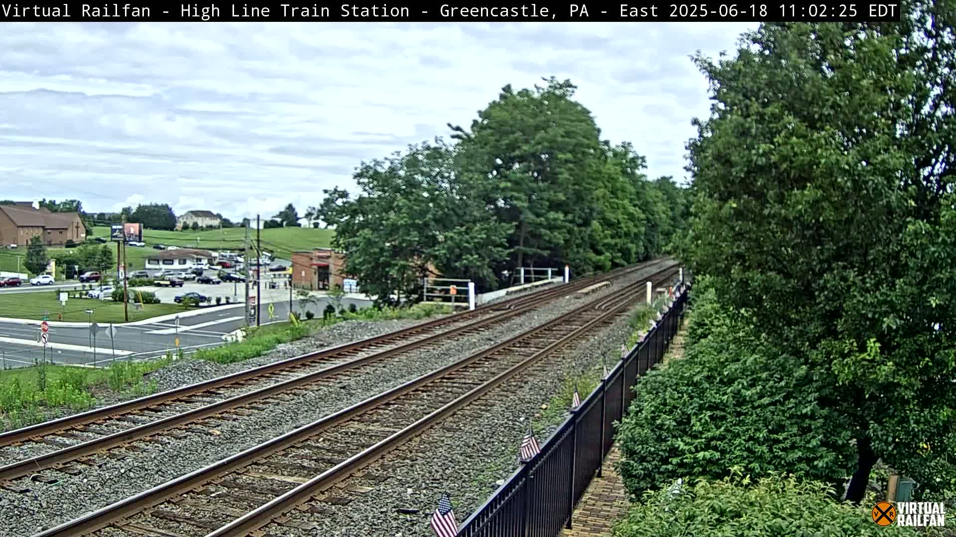 A view of a railway track stretching into the distance under a partly cloudy sky, passing by a parking lot and buildings before entering a wooded area.