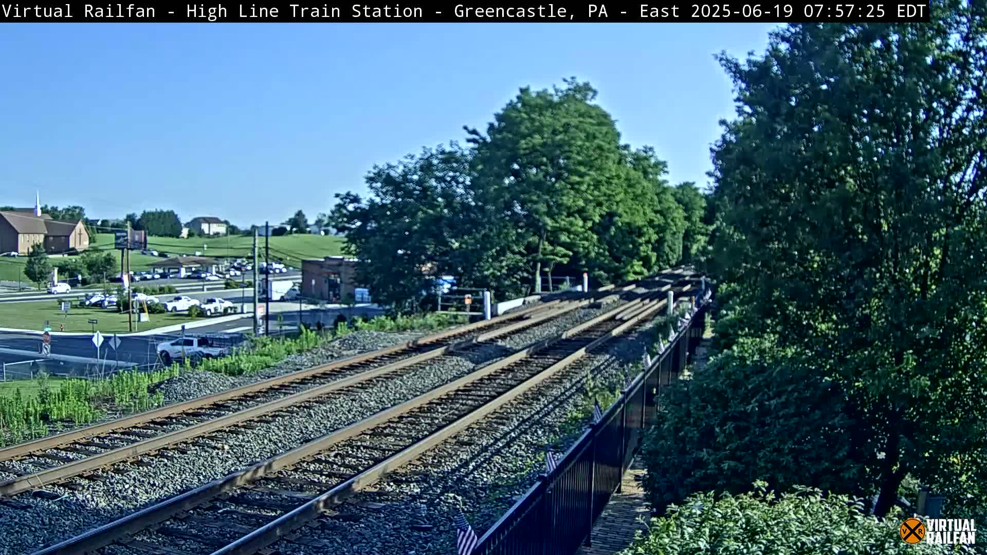 A high-angle view of three sets of train tracks on a sunny day, with lush green trees and a parking lot visible in the background.