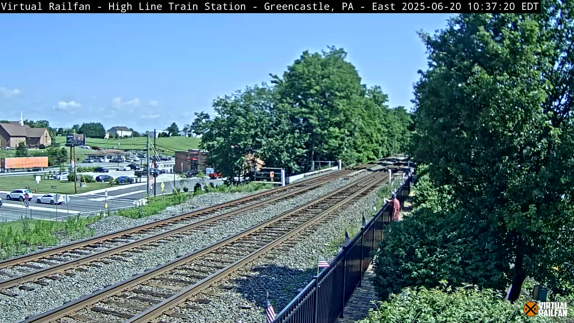 A high-angle view of train tracks on a sunny day, with a road and buildings visible in the background.