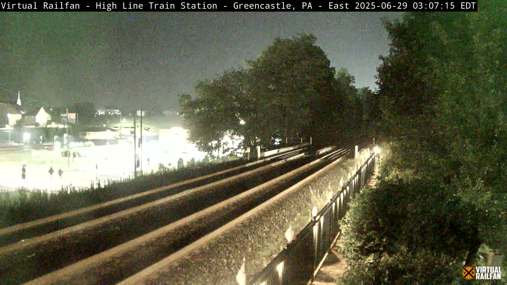 Three sets of train tracks are visible at night, bordered by lush green vegetation under a dark sky.