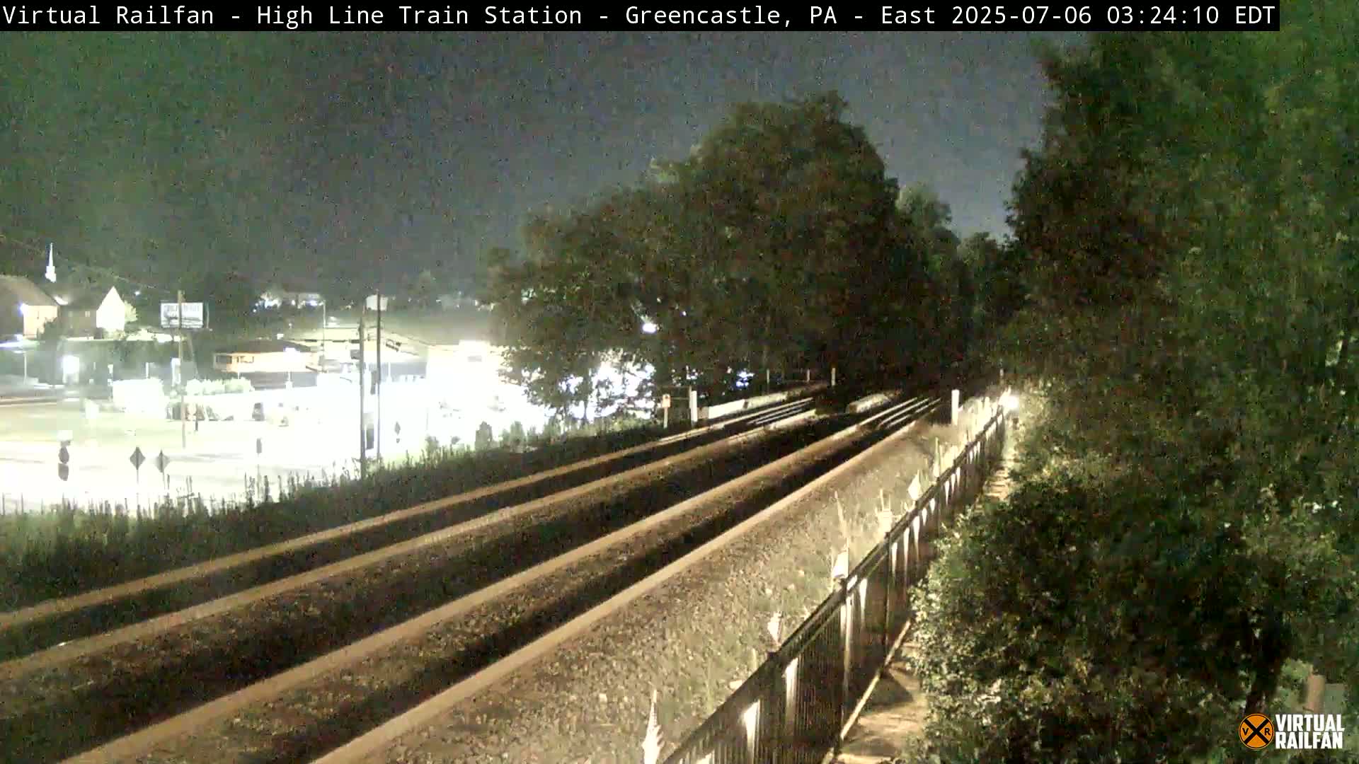 At night, empty train tracks extend into the distance, bordered by a fence and lush greenery under a dark sky.