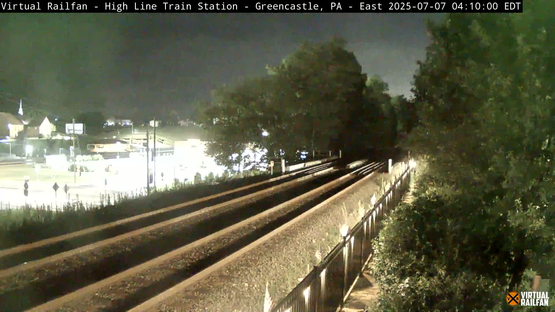 Here is a description of the image in a single sentence: At night, two sets of train tracks stretch into the distance, bordered by a fence and lush green vegetation, with a dimly lit road and buildings visible in the background.