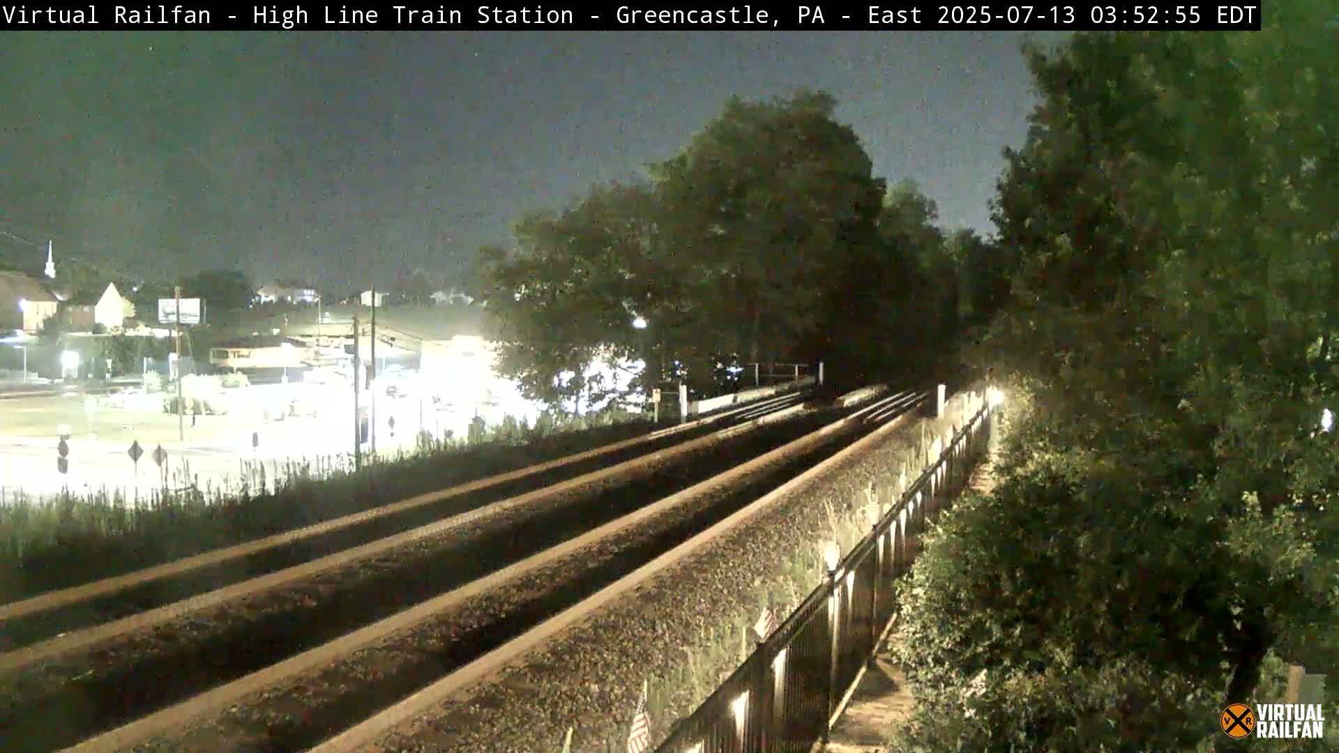 At night, two sets of train tracks are visible, bordered by vegetation and a fence, with a street and buildings in the background under a dark sky.