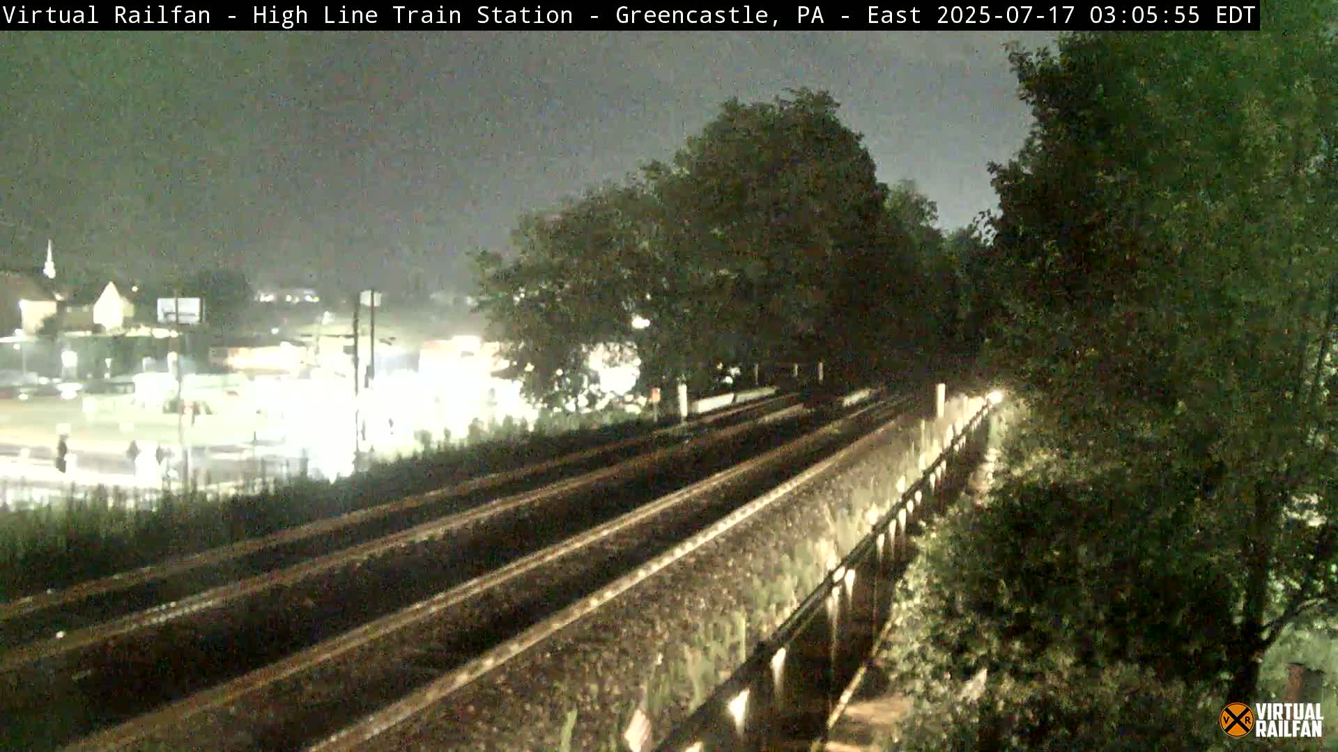 A nighttime view of train tracks on a bridge overlooking a dimly lit area, possibly a sports field, with lush green trees lining the sides of the bridge under a dark, overcast sky.