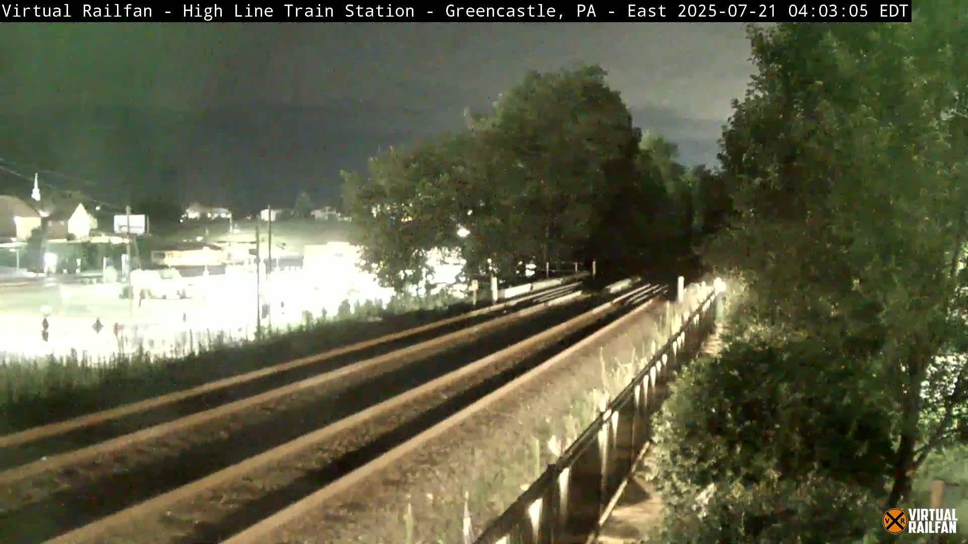 A nighttime view of a double-track railway line stretches into the distance, flanked by verdant trees under a dark sky.