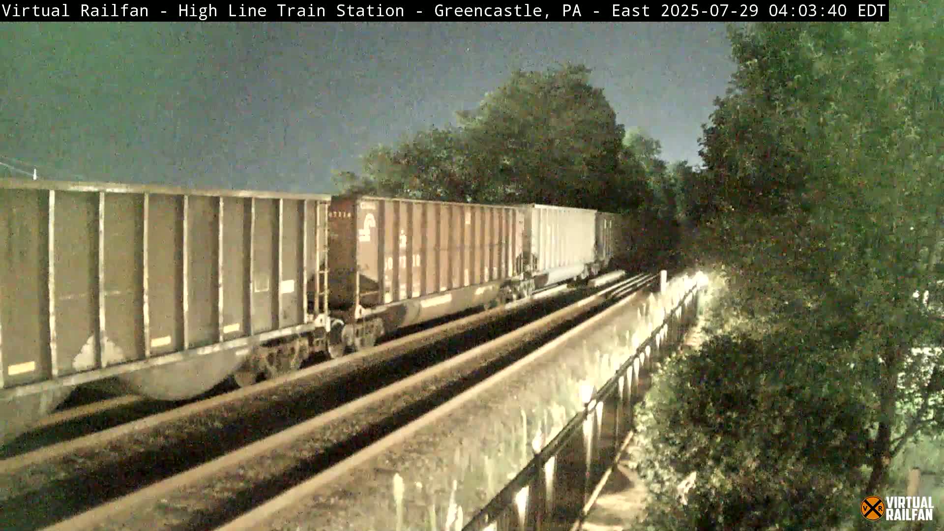 A nighttime view of a train of covered hopper cars moving along railroad tracks beside a tree-lined embankment.