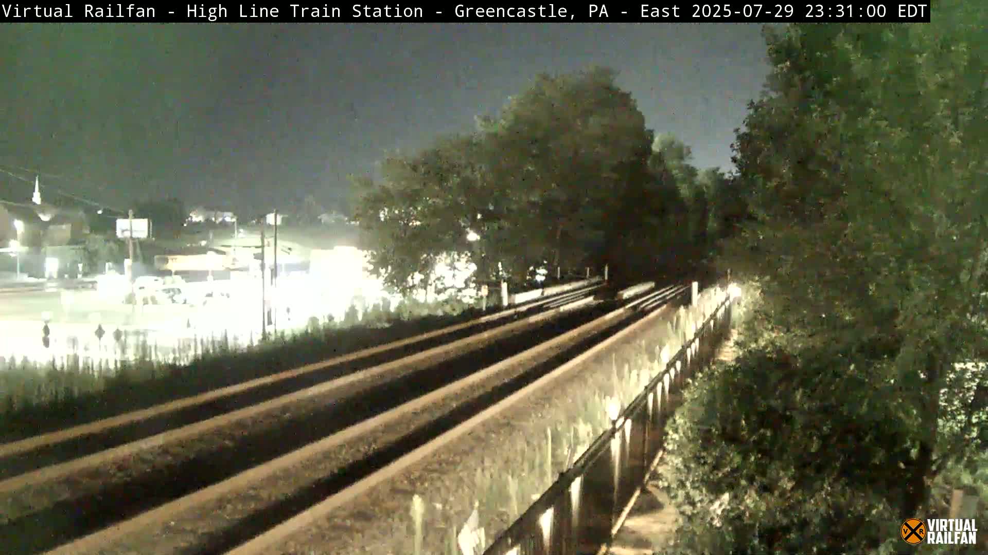 A nighttime view of a train of covered hopper cars moving along railroad tracks beside a tree-lined embankment.