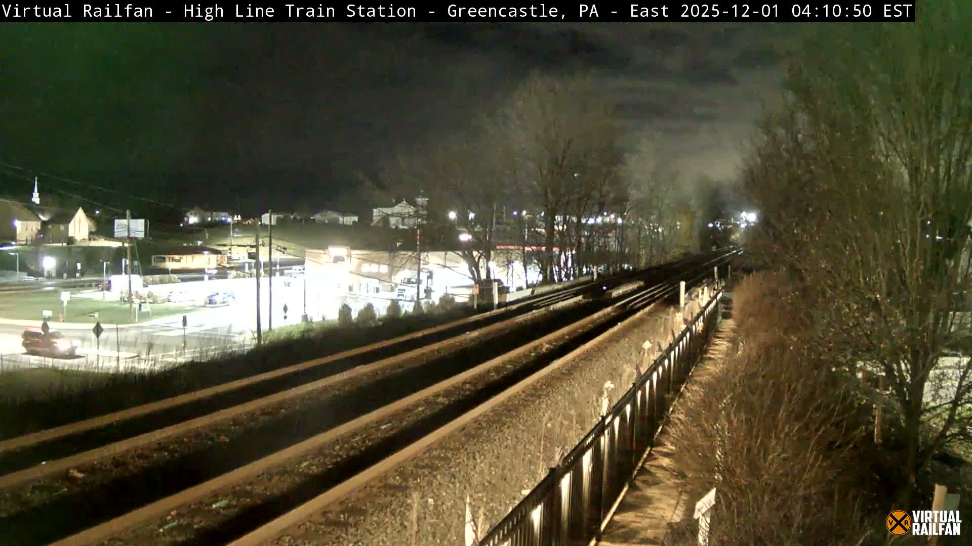 An outdoor nighttime scene displays multiple train tracks under a cloudy sky, with artificial lights illuminating a distant road intersection, buildings, and trees.