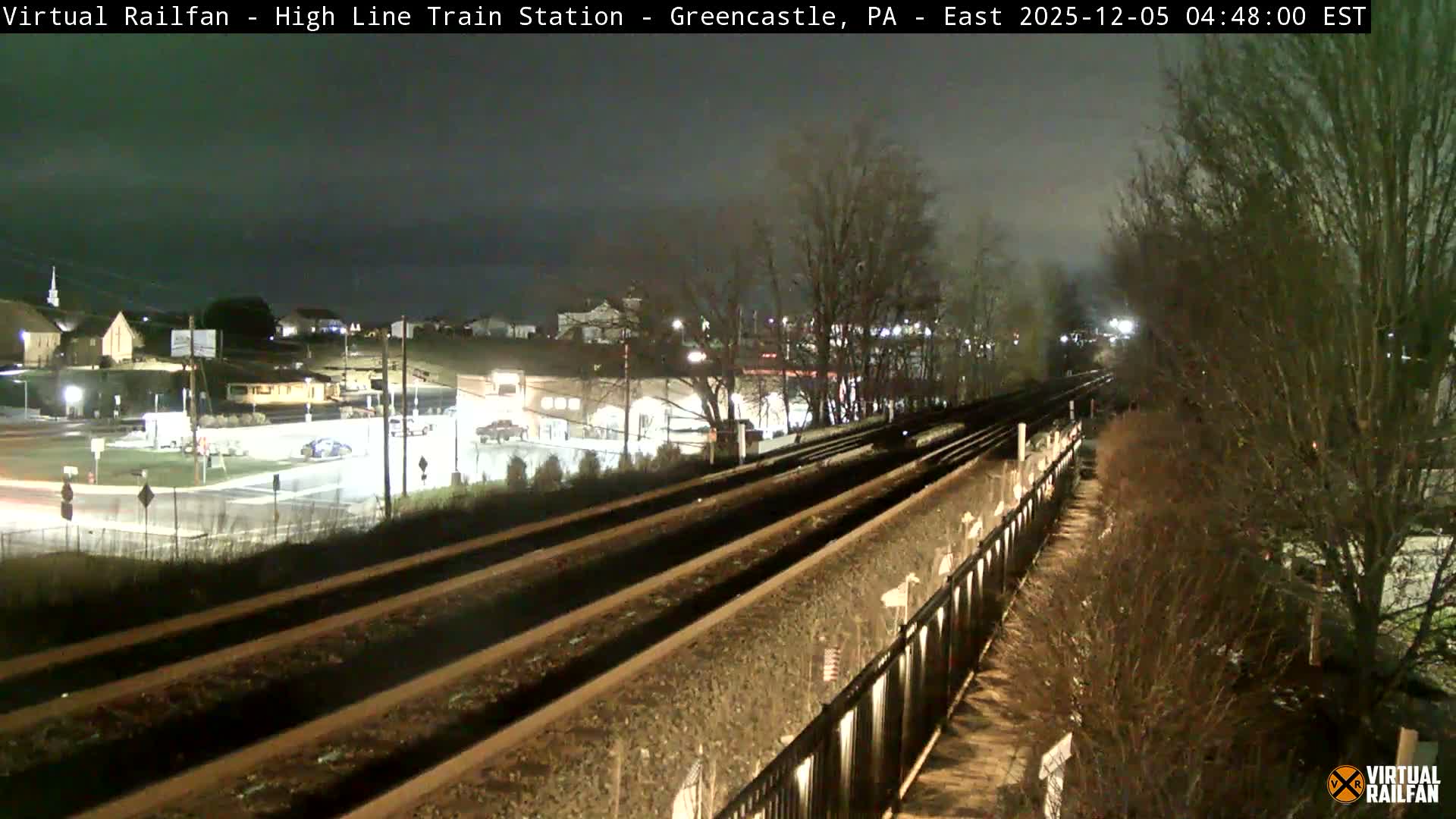 An early morning scene captures multiple train tracks running diagonally through a residential and commercial area with illuminated buildings and an intersection on the left, flanked by bare trees and bushes on the right, all under a dark, overcast sky.