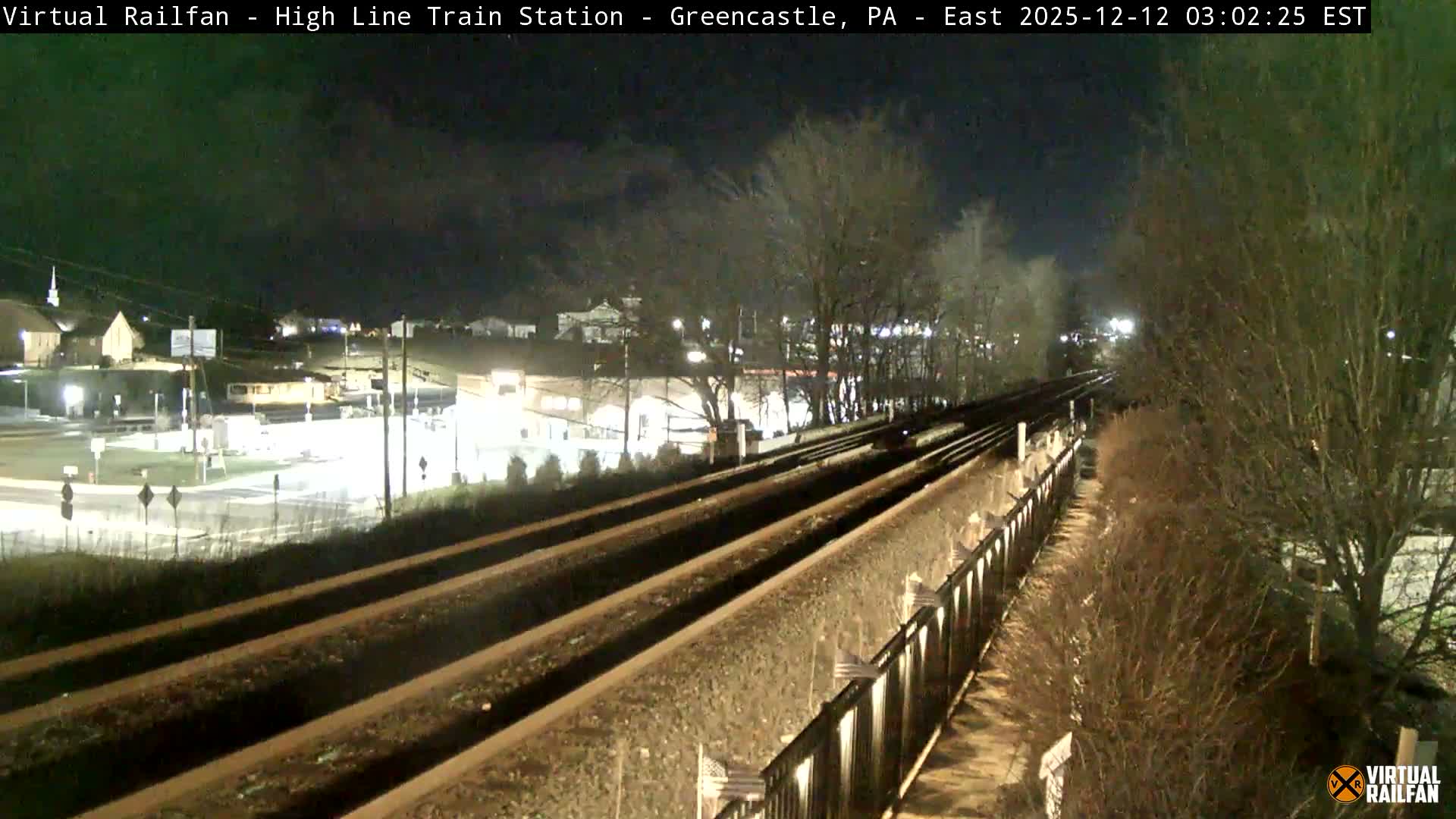 An elevated nighttime view shows multiple railroad tracks curving into the distance, with a brightly lit town featuring buildings and roads on the left, and bare winter trees and a retaining wall lining the tracks on the right, all under a clear or partly cloudy sky with no visible precipitation.