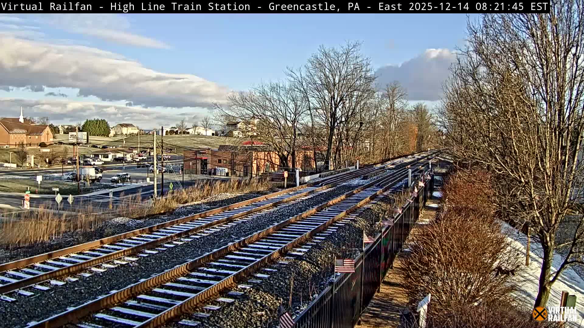 An elevated nighttime view shows multiple railroad tracks curving into the distance, with a brightly lit town featuring buildings and roads on the left, and bare winter trees and a retaining wall lining the tracks on the right, all under a clear or partly cloudy sky with no visible precipitation.