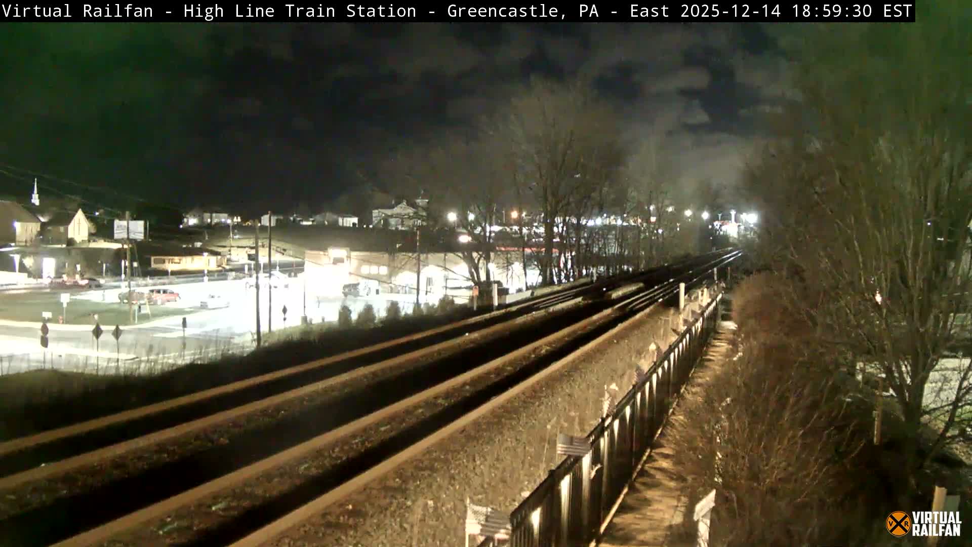 An elevated nighttime view shows multiple railroad tracks curving into the distance, with a brightly lit town featuring buildings and roads on the left, and bare winter trees and a retaining wall lining the tracks on the right, all under a clear or partly cloudy sky with no visible precipitation.
