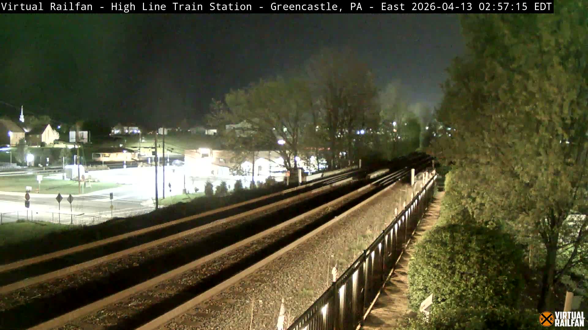 An elevated nighttime view shows multiple railroad tracks curving into the distance, with a brightly lit town featuring buildings and roads on the left, and bare winter trees and a retaining wall lining the tracks on the right, all under a clear or partly cloudy sky with no visible precipitation.