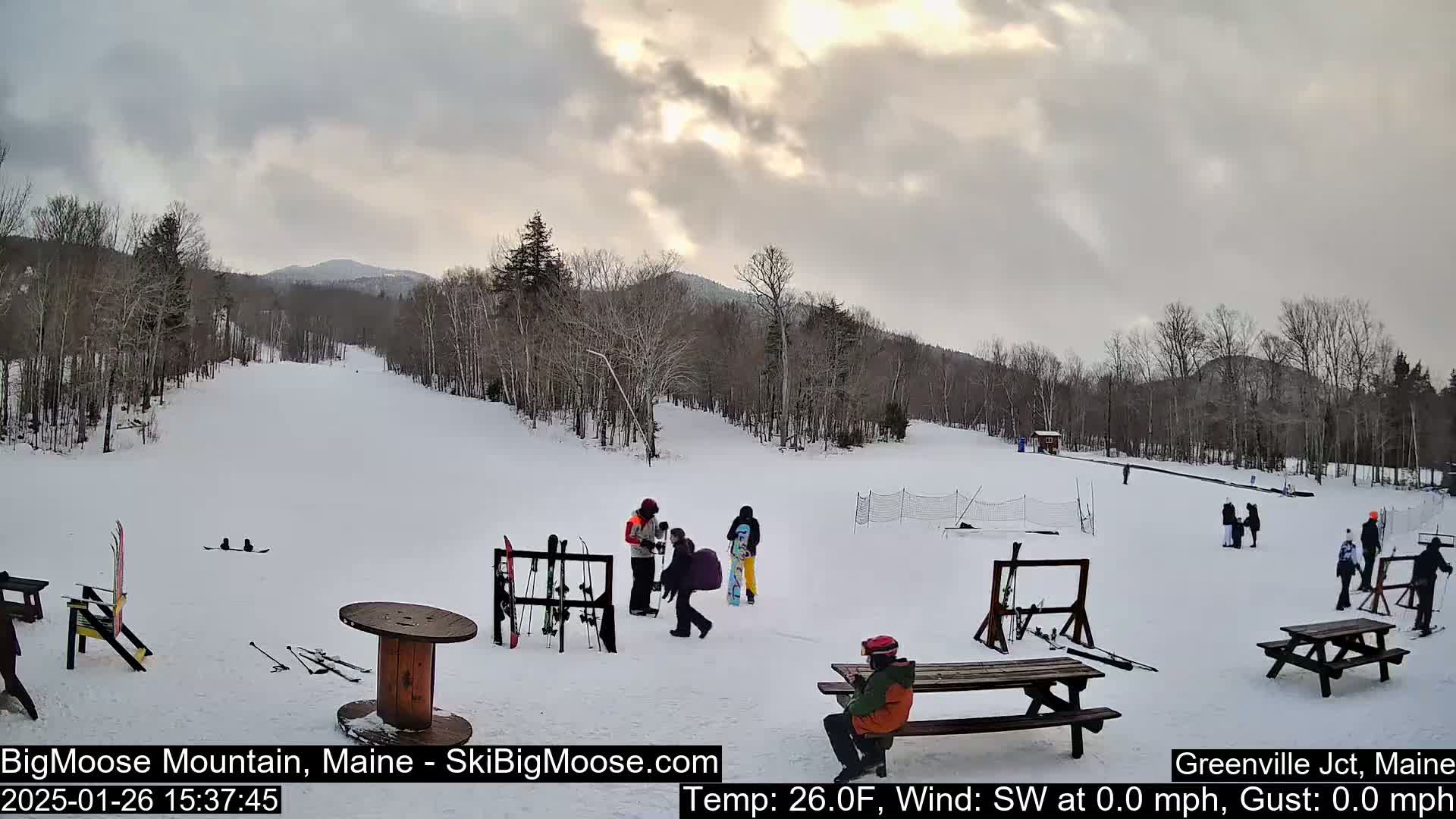 A snowy ski slope with several people gathered near picnic tables and ski racks under a cloudy sky.