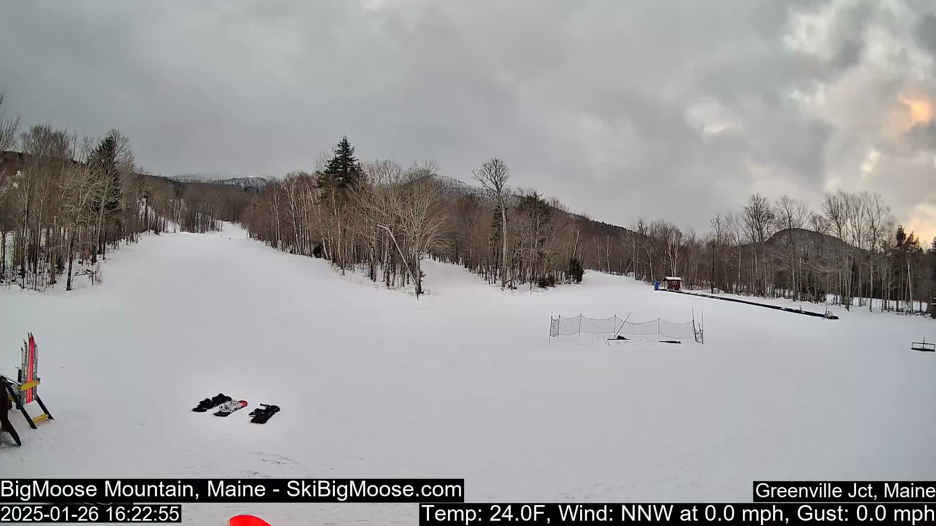A snow-covered ski slope is seen under an overcast sky, with bare trees lining the sides and a small building visible in the distance.