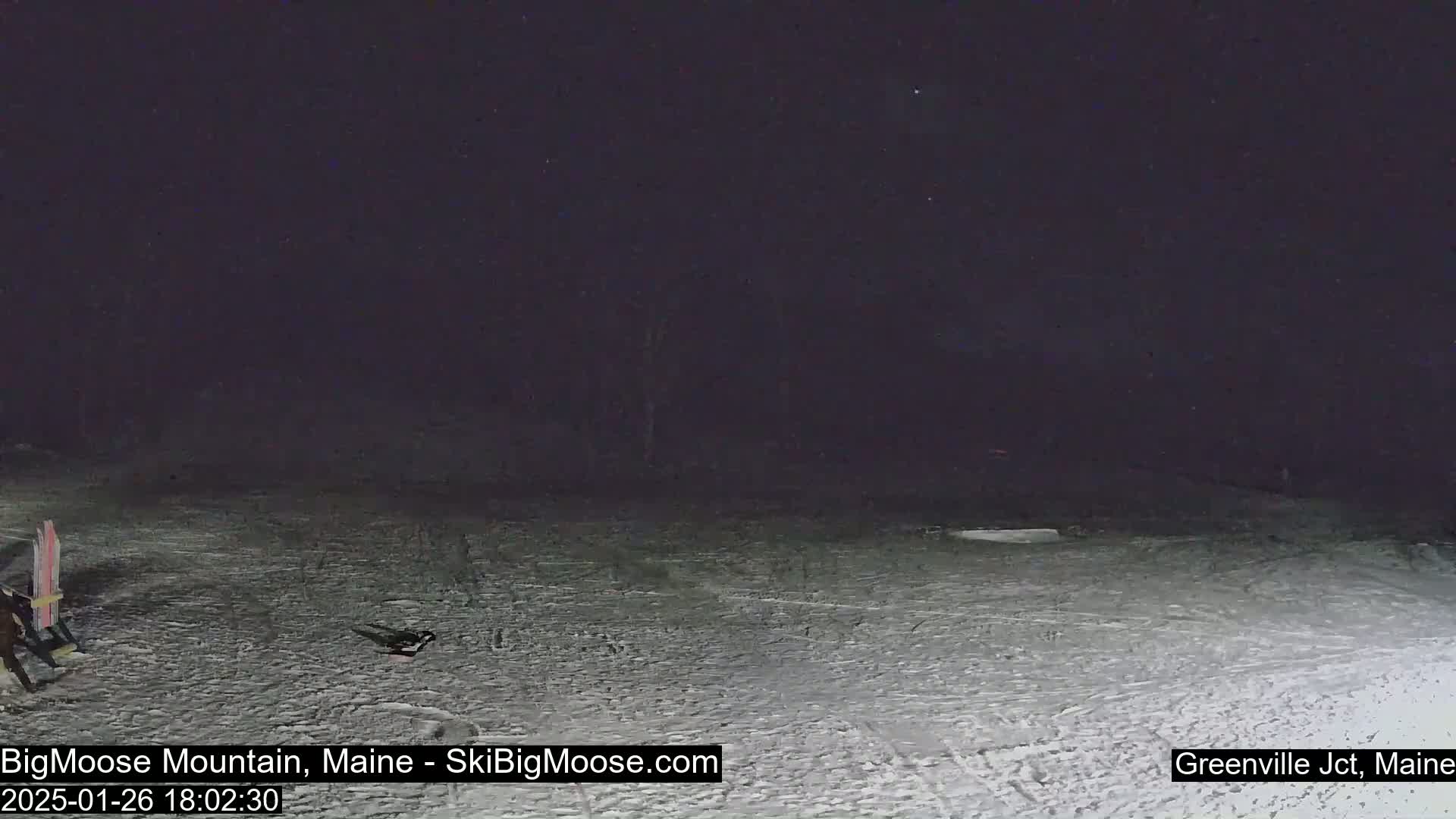 The image shows a snow-covered area at night, with a person and some skis visible in the lower left corner.