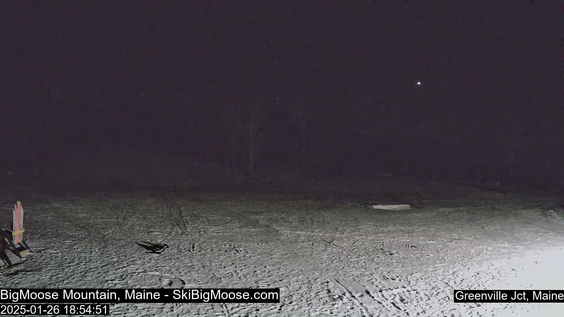 A nighttime, snow-covered scene shows a mostly empty snow-covered slope with a few people visible near some skis and a chair.