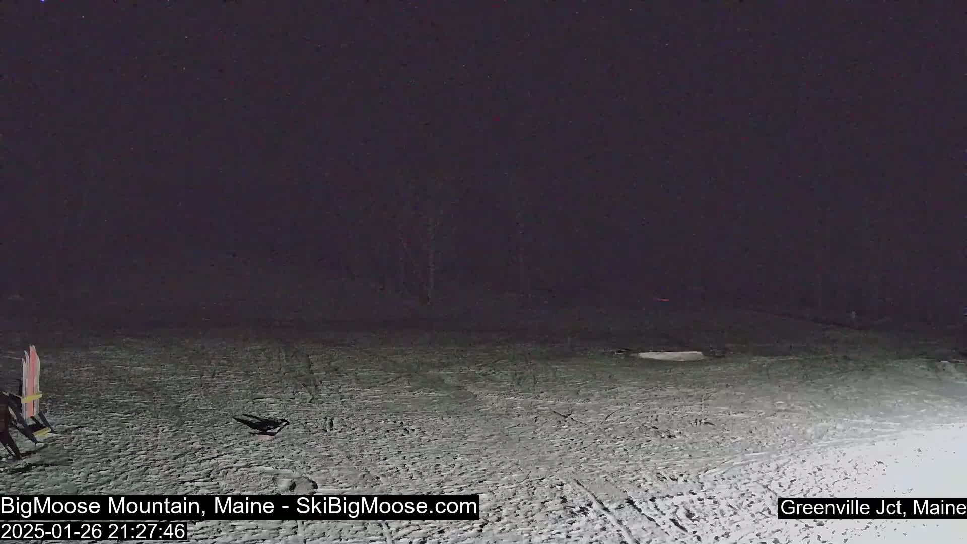 The image shows a snow-covered area at night, with a pair of skis leaning against something in the bottom-left corner.