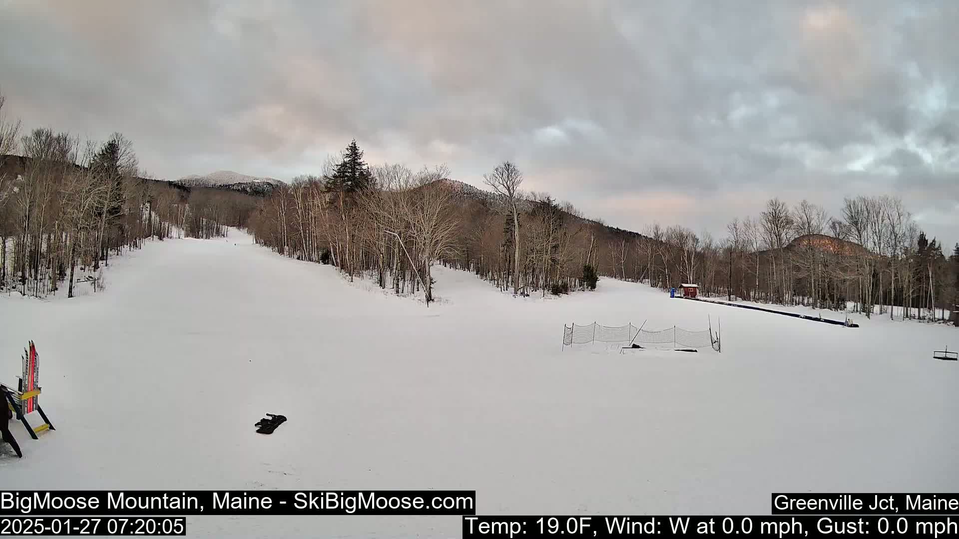 A snow-covered ski slope is visible beneath a mostly cloudy sky, with bare deciduous trees lining the hills in the background.