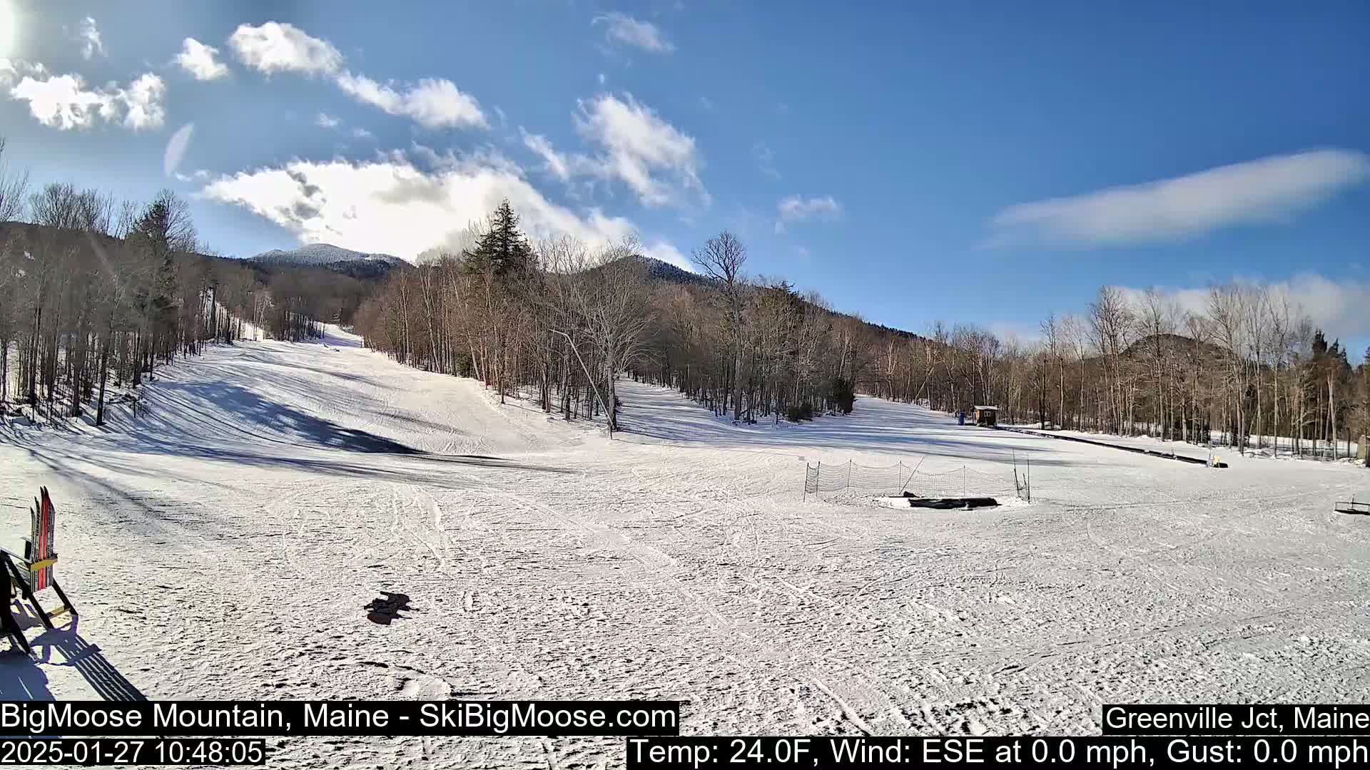A snow-covered ski slope with trees and mountains in the background under a mostly sunny sky.
