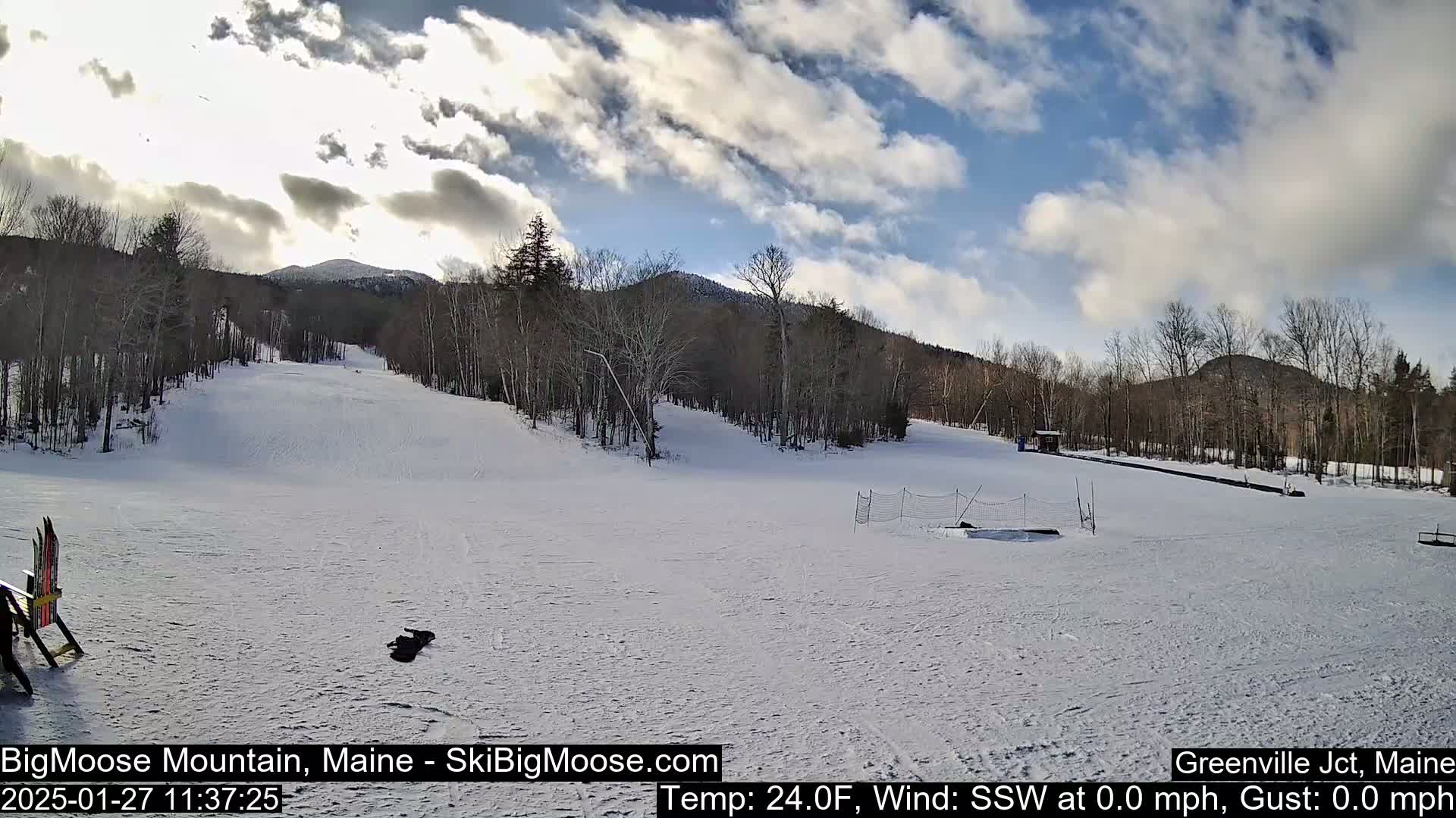A snow-covered ski slope with trees and mountains in the background under a partly cloudy sky.