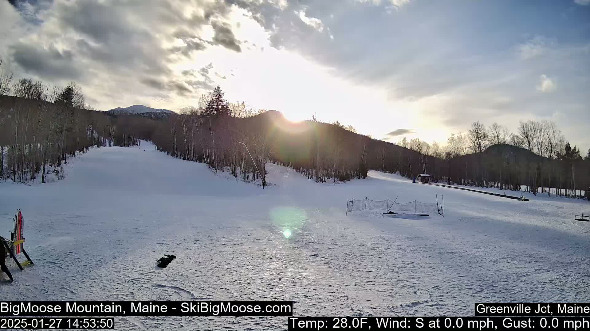A snow-covered ski slope is bathed in the bright light of a setting sun under partly cloudy skies.