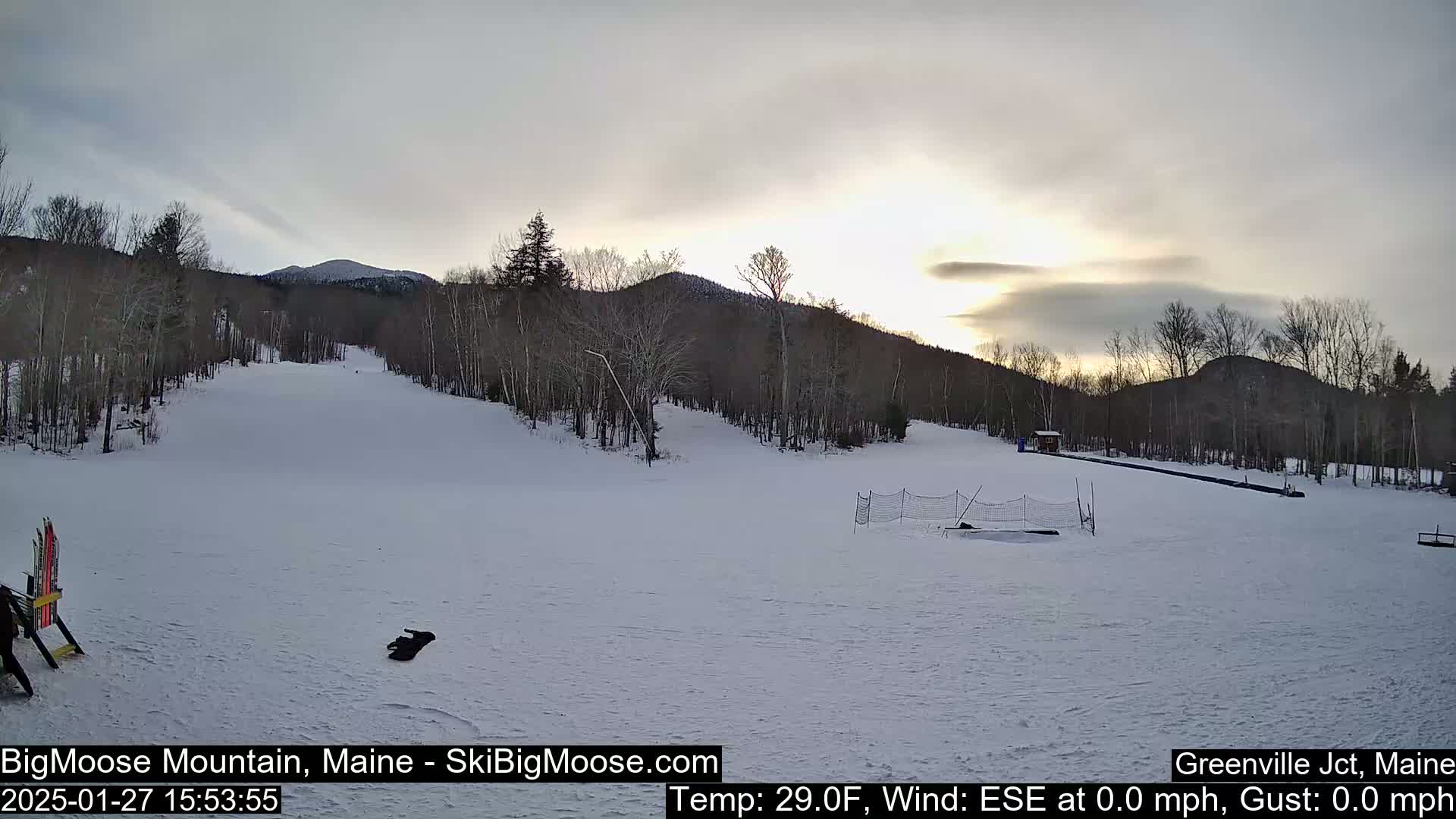 A snow-covered ski slope at sunset, with bare trees and mountains in the background under a cloudy sky.