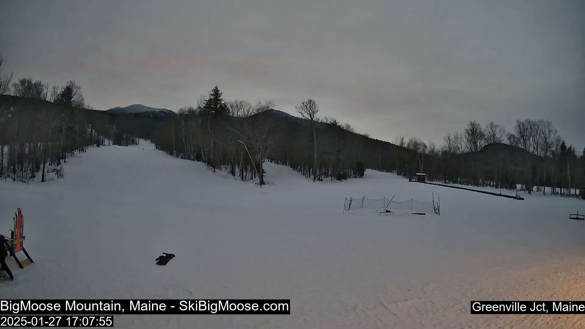A snow-covered ski slope is visible under an overcast sky, with trees lining the background and a small building in the distance.