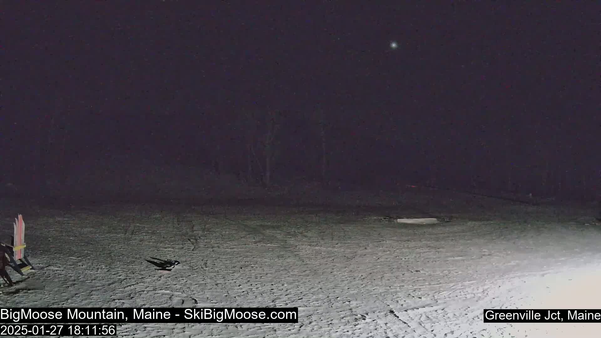 A snow-covered, dark area at night with a few objects and faint tracks in the snow, under a clear sky.