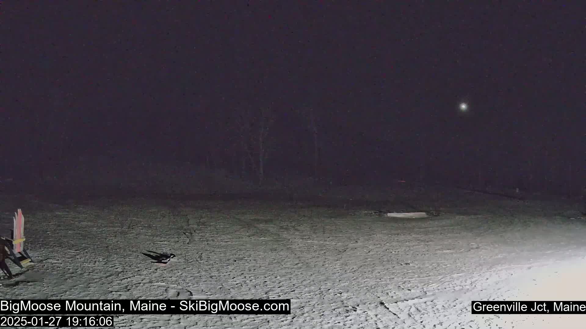 A snow-covered area at night with a bright light visible in the dark sky and some skis leaning against what appears to be a chair.