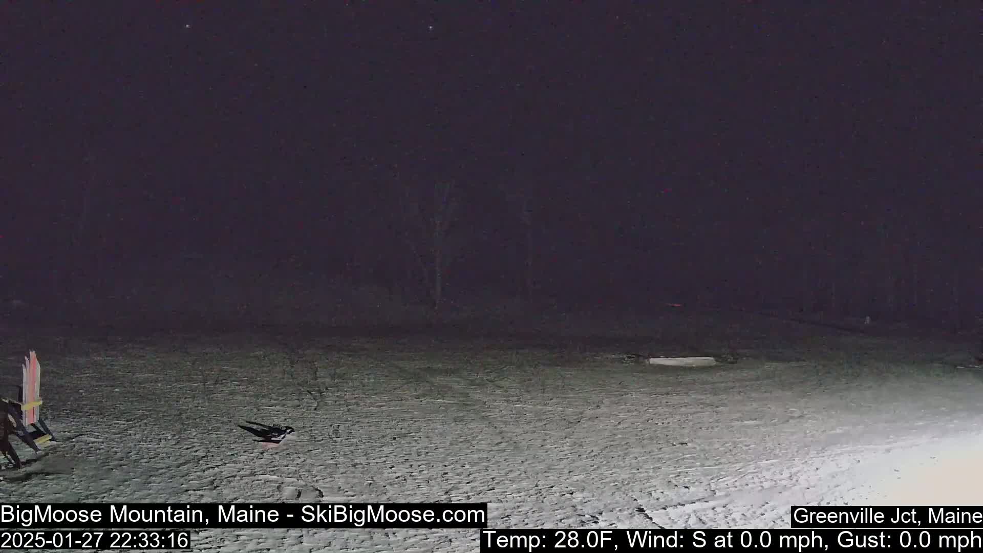 A snow-covered, dark nighttime scene shows a mostly empty area with a lone chair and skis to the left, under a clear, starlit sky.