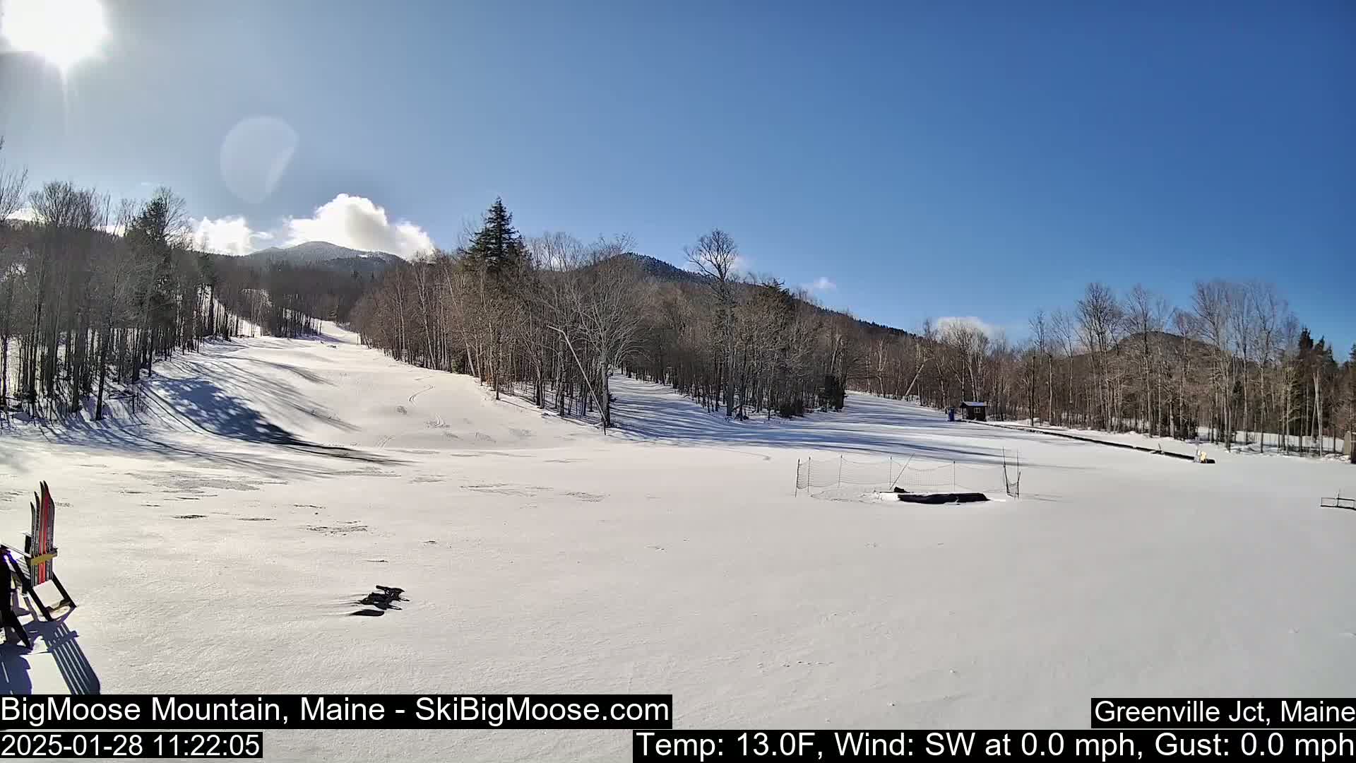 A snow-covered ski slope with trees and mountains in the background under a mostly sunny sky.