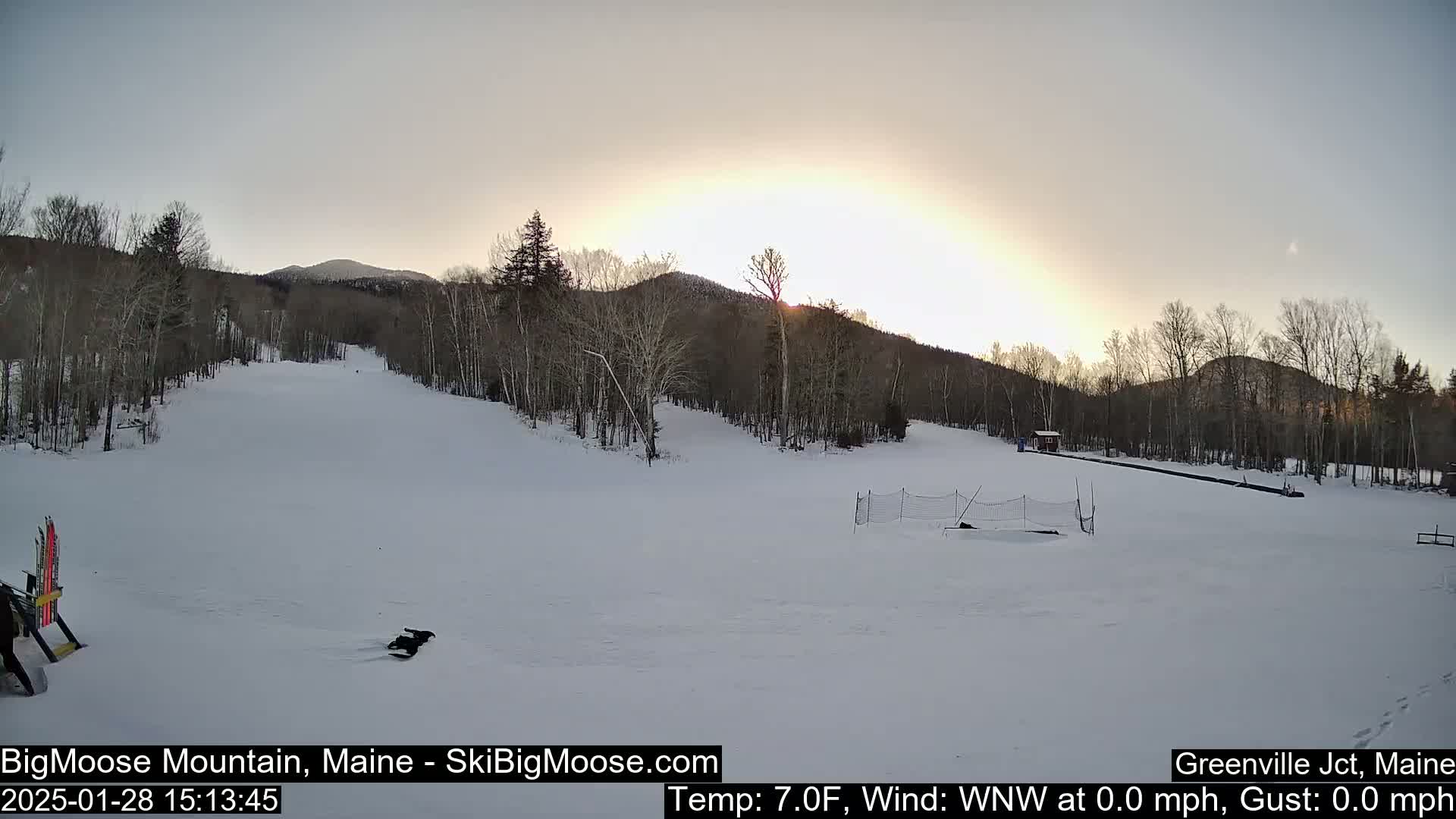 A snow-covered ski slope is bathed in the soft light of a setting sun, with bare trees lining the hills in the background.