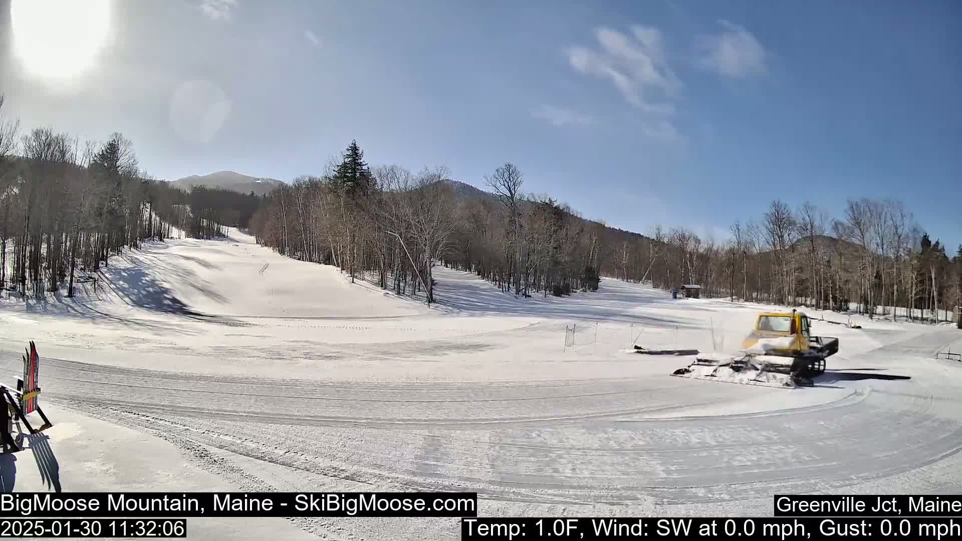 A snowcat grooms a snow-covered ski slope under a sunny sky, with bare deciduous trees lining the hills in the background.