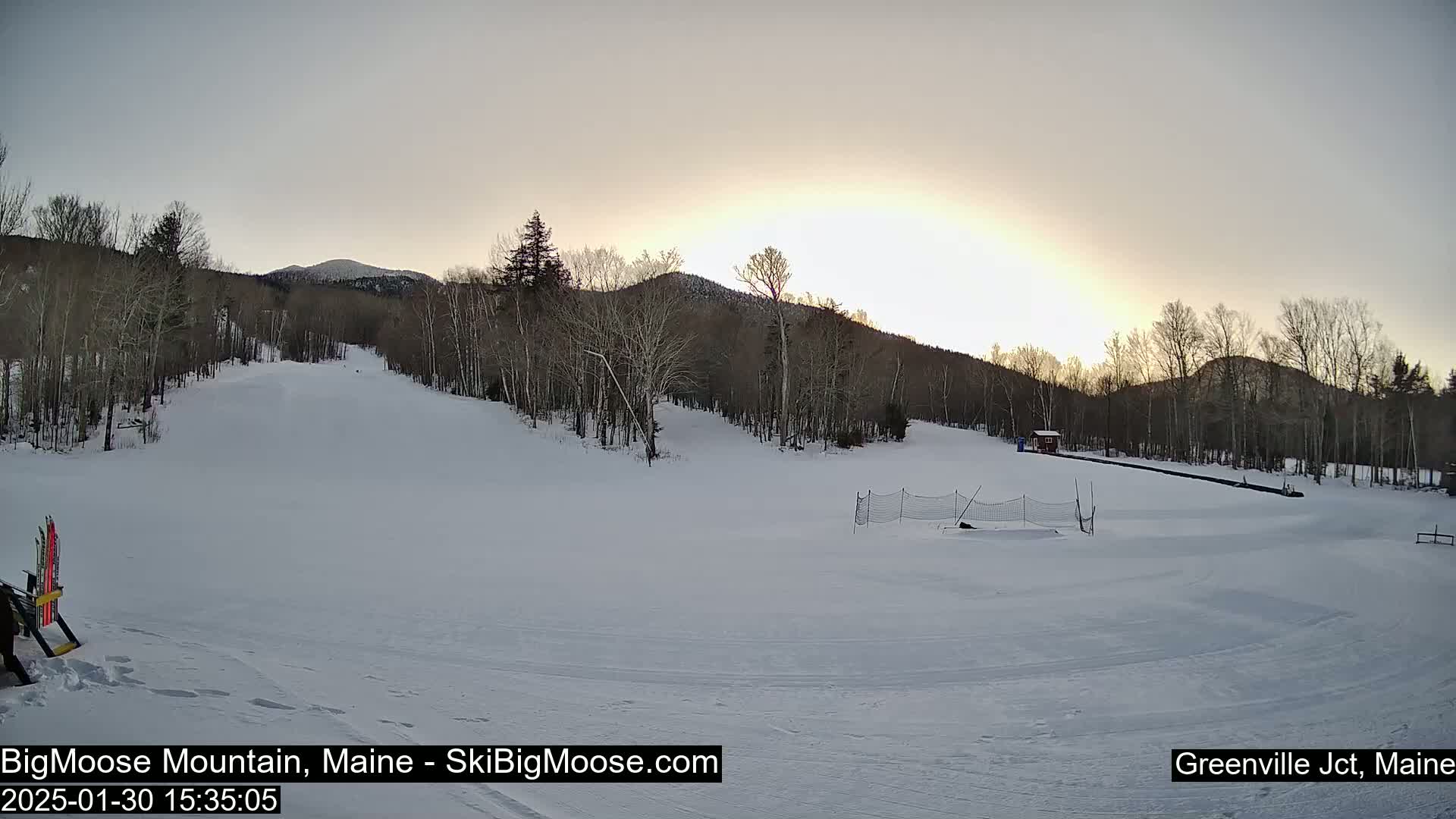 A snow-covered ski slope with trees and mountains in the background under a bright, sunset sky.