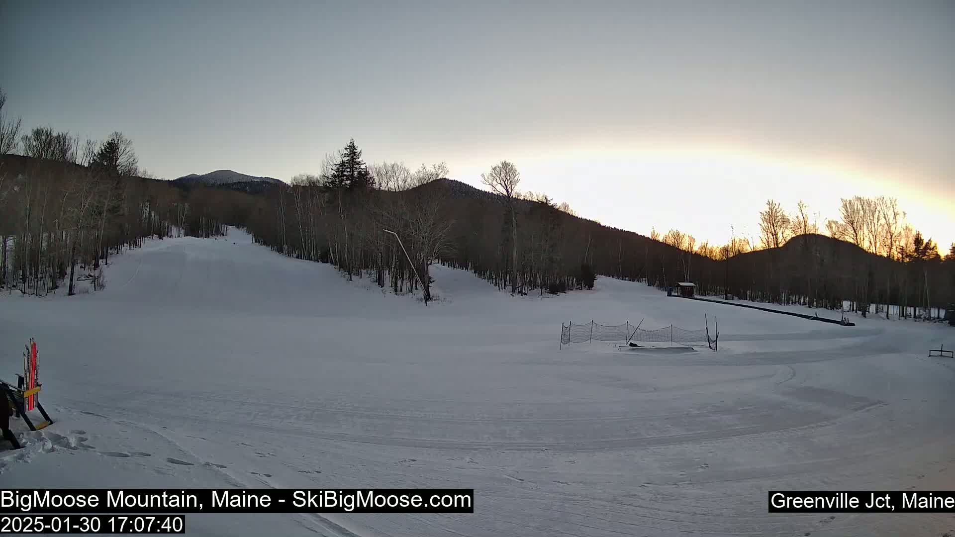 A snow-covered ski slope at sunset is surrounded by leafless trees and mountains.
