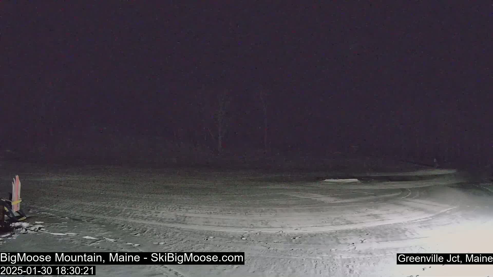 A snow-covered, dark area with tire tracks and a pair of skis visible in the lower left corner under a nighttime sky.