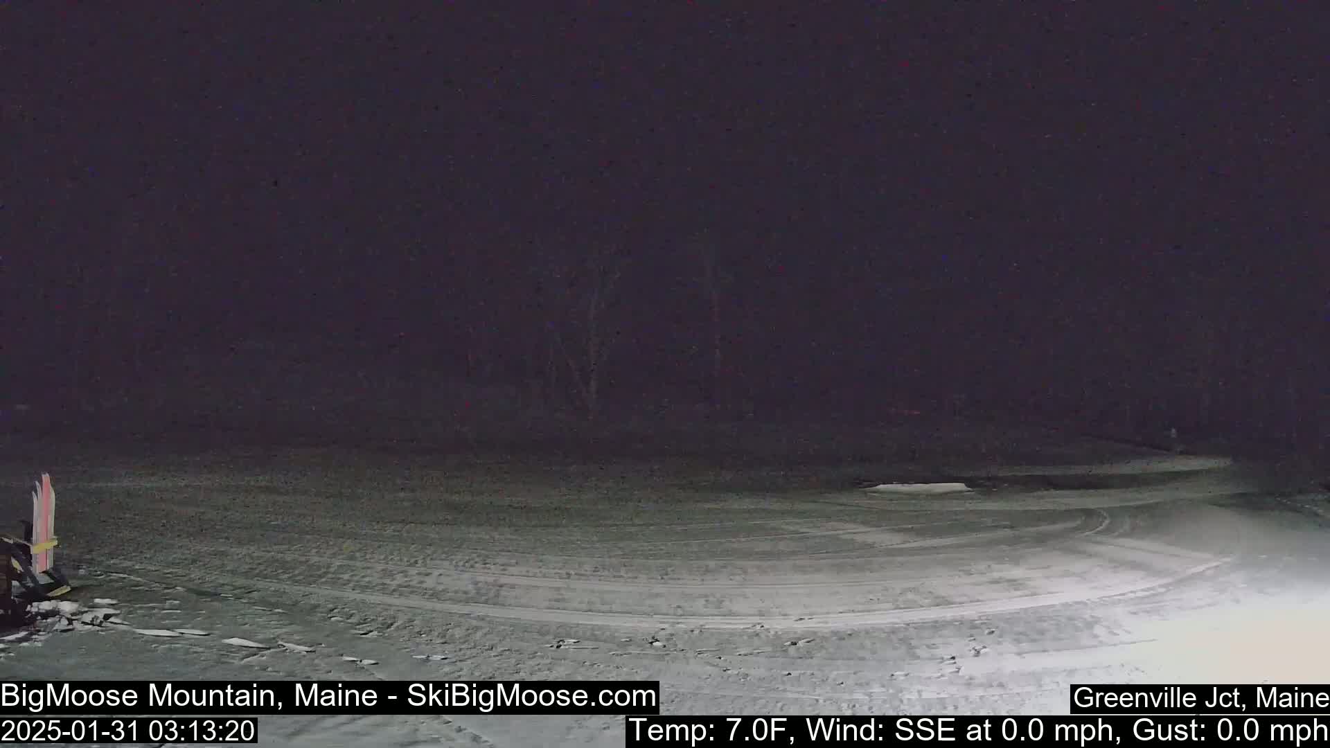 A snow-covered, dark nighttime scene shows tire tracks curving across a flat, empty area, with a few dark trees in the background and skis leaning against an object to the lower left.