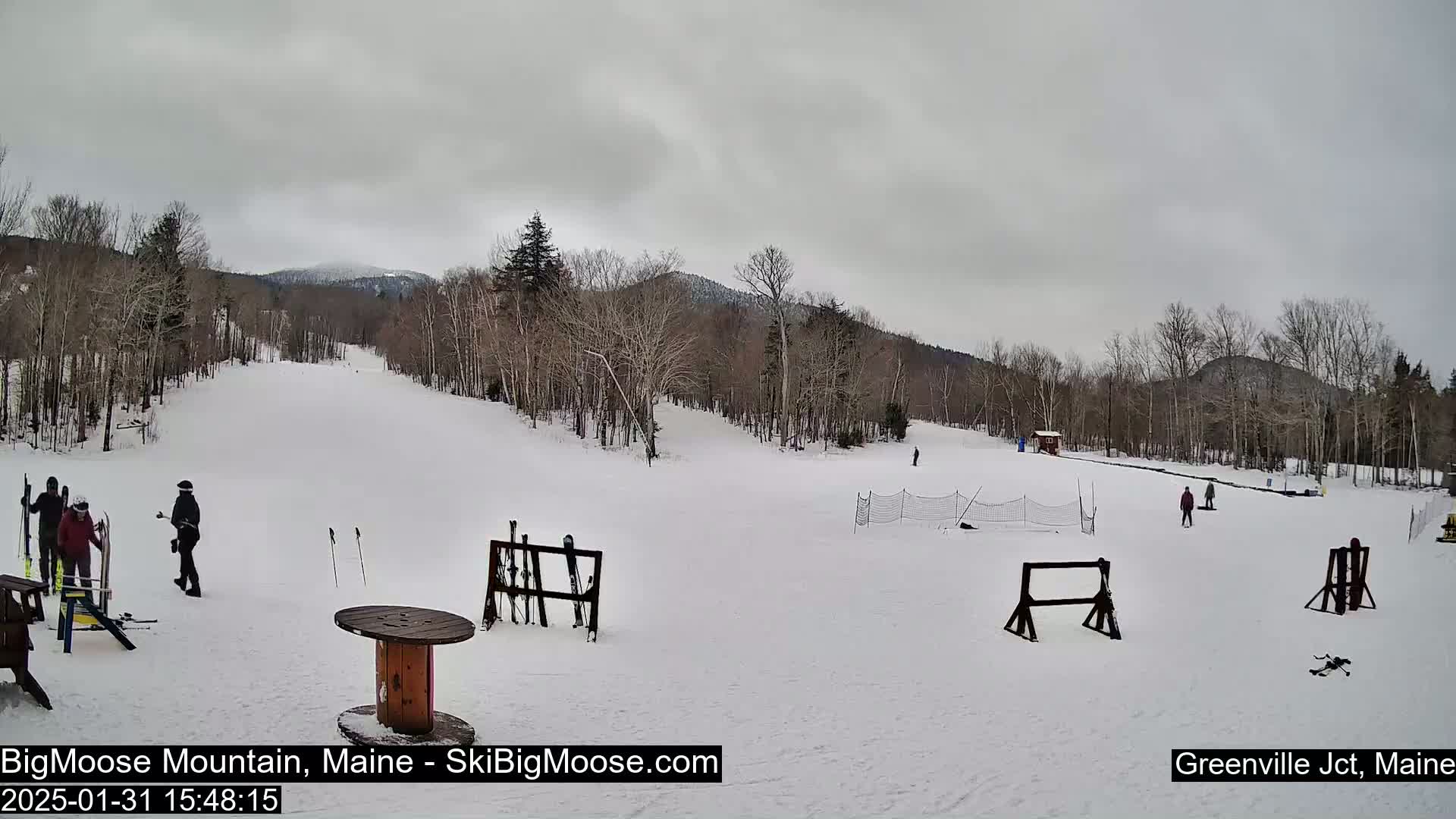 A snow-covered ski slope with several people skiing and several ski racks is visible under an overcast sky.