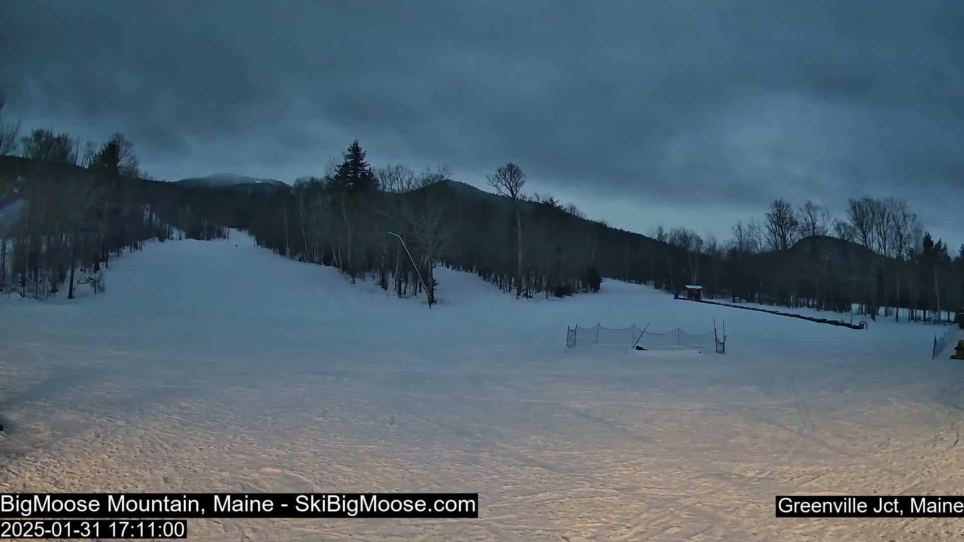 A snow-covered ski slope is visible under a dark, cloudy sky at dusk, with trees lining the hills in the background.