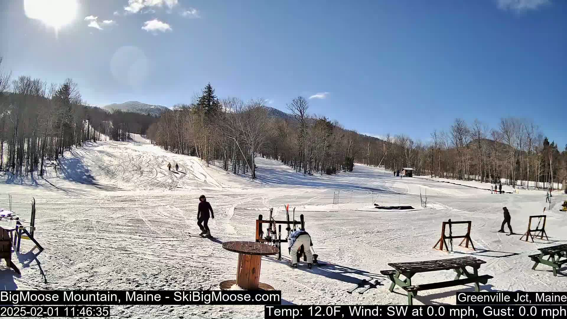 A sunny day at a snow-covered ski resort shows several skiers and snowboarders on the slopes, with mountains in the background.
