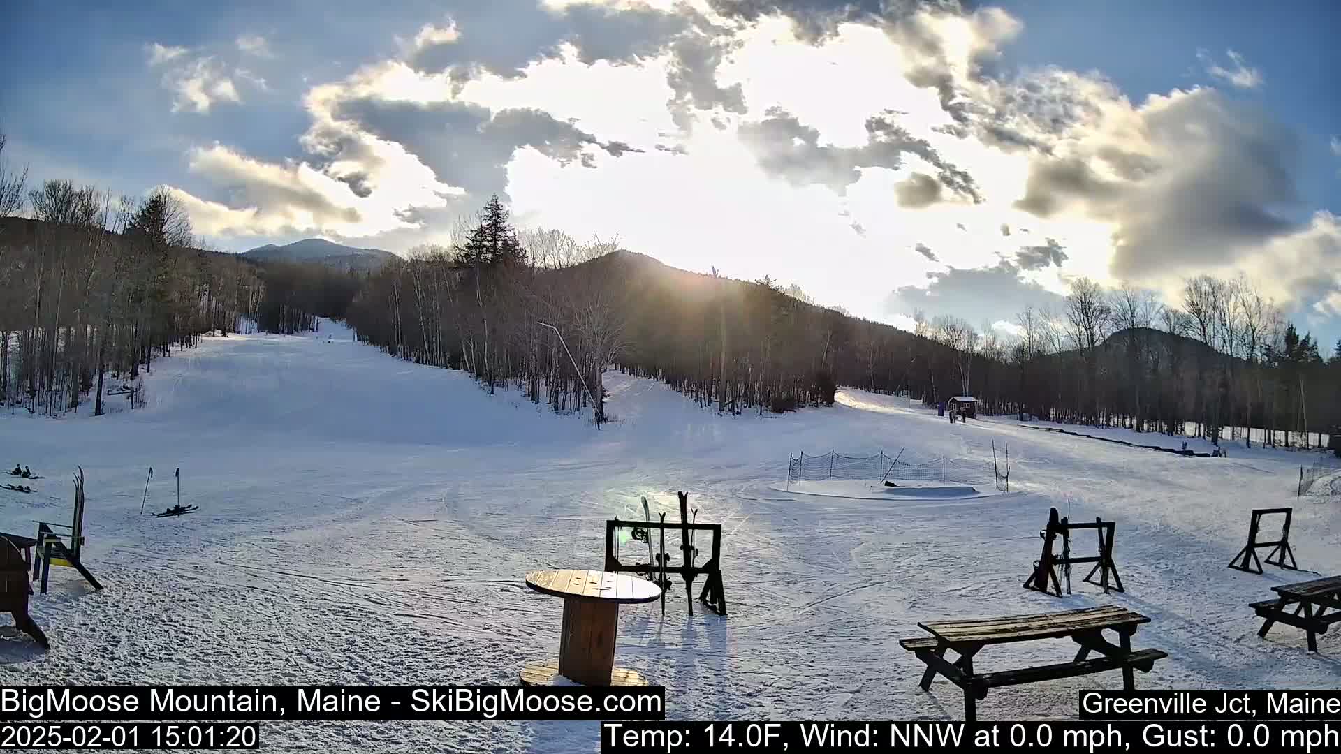 A snow-covered ski slope is visible under a partly cloudy sky, with the sun shining through the clouds.