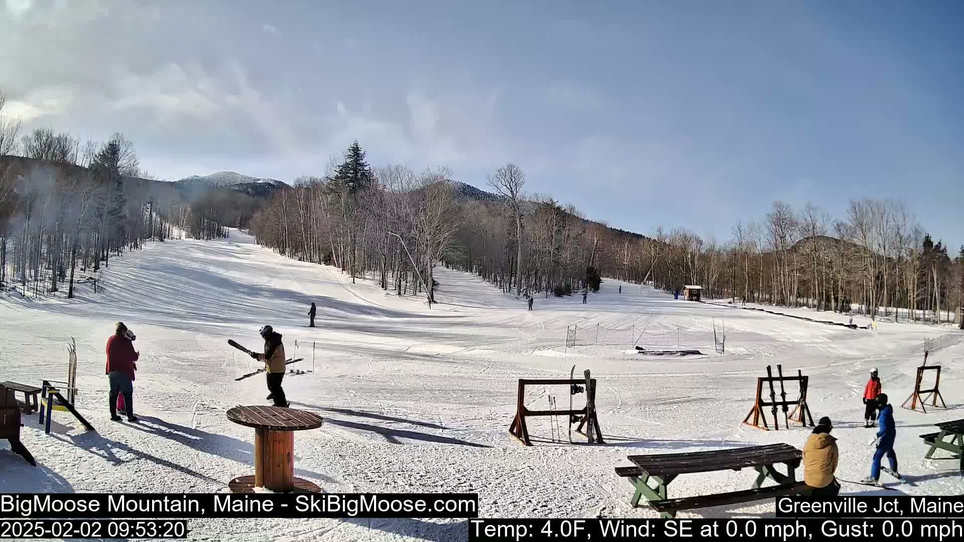 A snowy ski slope with several people skiing and standing around, under a mostly sunny sky.