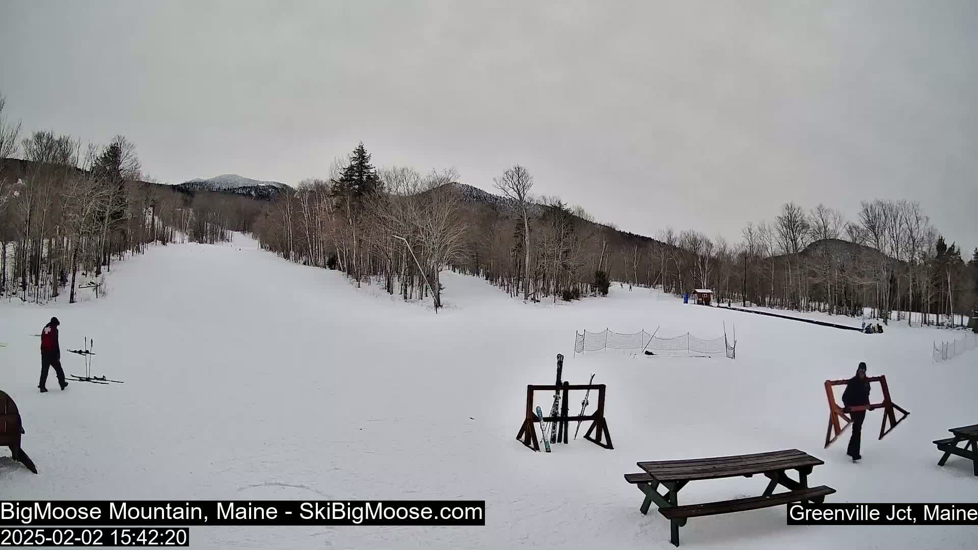 A snow-covered ski slope is visible with several people, some carrying skis, under an overcast sky.